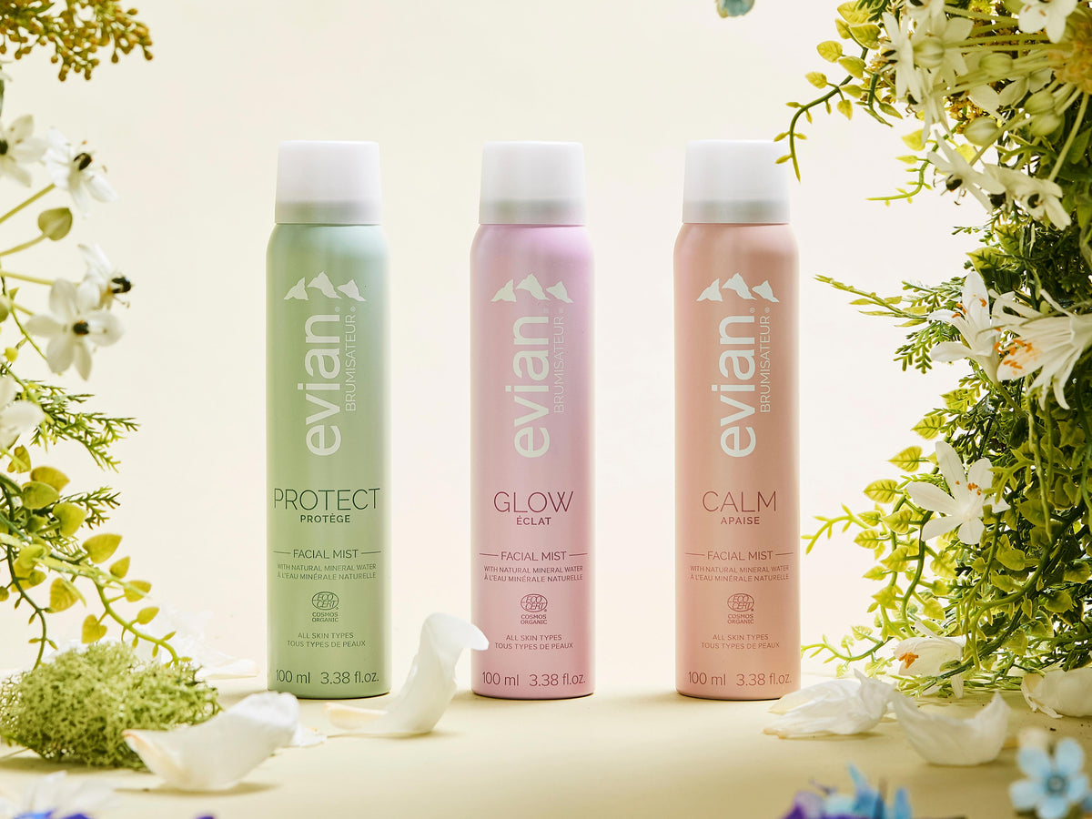 Evian Protect, Glow and Calm facial mists in pastel green, pink and peach cans, surrounded by white flowers and greenery on a pale background