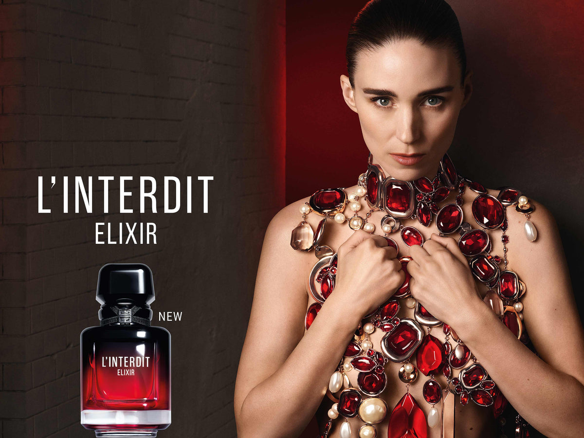 Model in an ornate top of red jewels and pearls beside the Givenchy L'Interdit Elixir bottle, black glass fading to red, with the words L'Interdit Elixir on a dark red and black backdrop