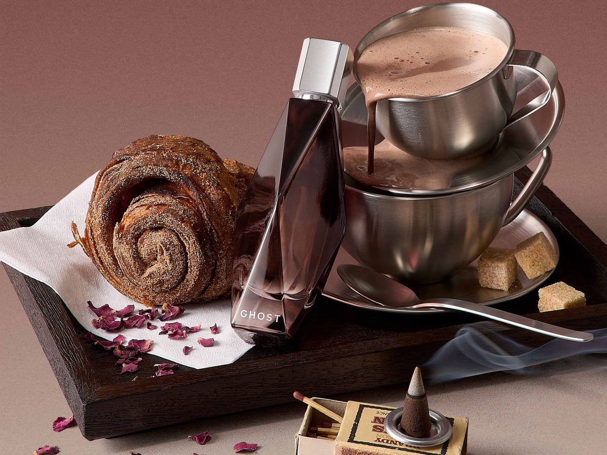 Ghost Moody Dream eau de parfum in a faceted smoky bottle, surrounded by stacked silver cups of hot chocolate, a cinnamon swirl pastry, sugar cubes, dried rose petals and incense on a wooden tray
