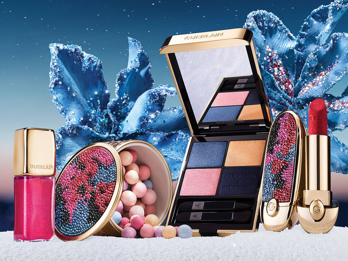 A luxury cosmetics set from Guerlain, including a lipstick, eyeshadow palette, lip gloss, and compact, styled with decorative blue crystals and pastel beads on a snowy surface.