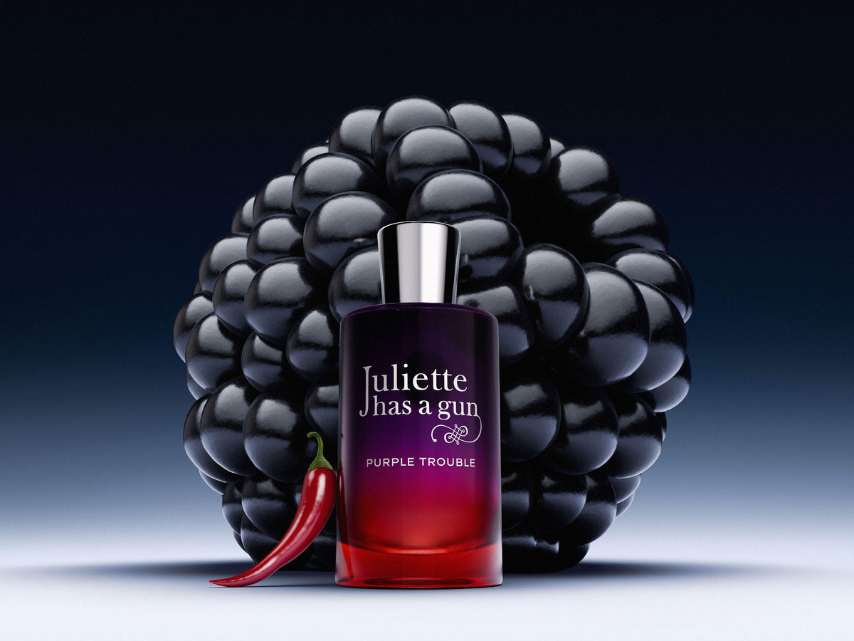 Juliette Has a Gun Purple Trouble perfume bottle in front of a large glossy black raspberry, with a red chili pepper at its base, on a dark blue gradient background.