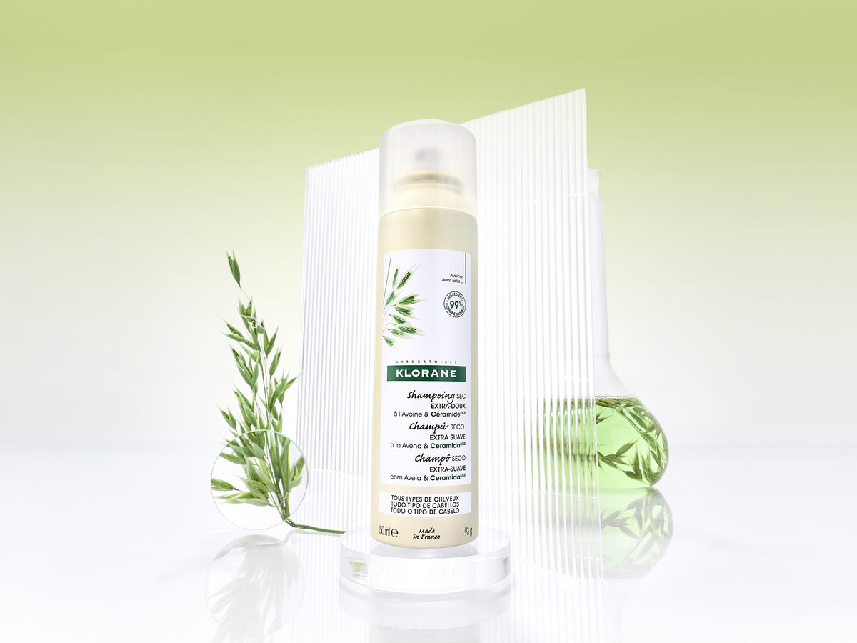 Klorane shampoo with olive in a white spray can with green label, beside a sprig of olive, a green glass flask and a ribbed glass panel on a pale green background