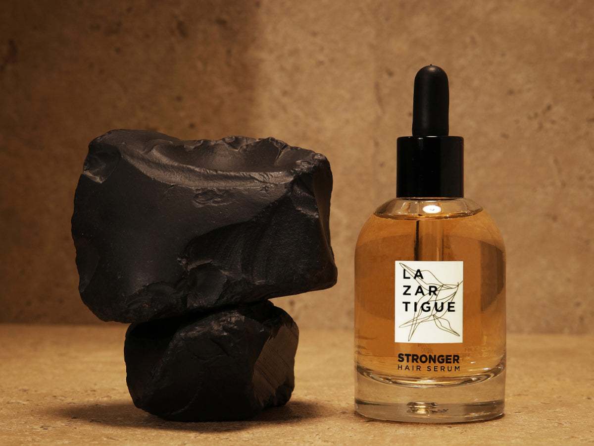 Lazartigue Stronger Hair Serum in a clear glass dropper bottle, beside stacked black stones on a warm sandy background