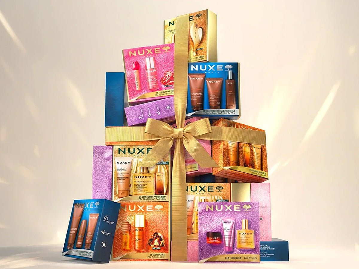 Tall tower of stacked Nuxe Christmas gift sets in pink, purple, blue and orange boxes, tied with a gold ribbon bow against a warm cream background