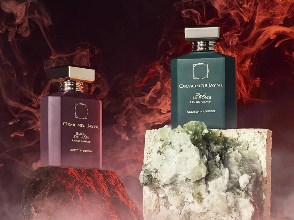Ormonde Jayne Bijou Zafran eau de parfum in a plum bottle and Oud Liaisons eau de parfum in a dark green bottle, both with gold caps, displayed on rough stone plinths amid swirling red smoke