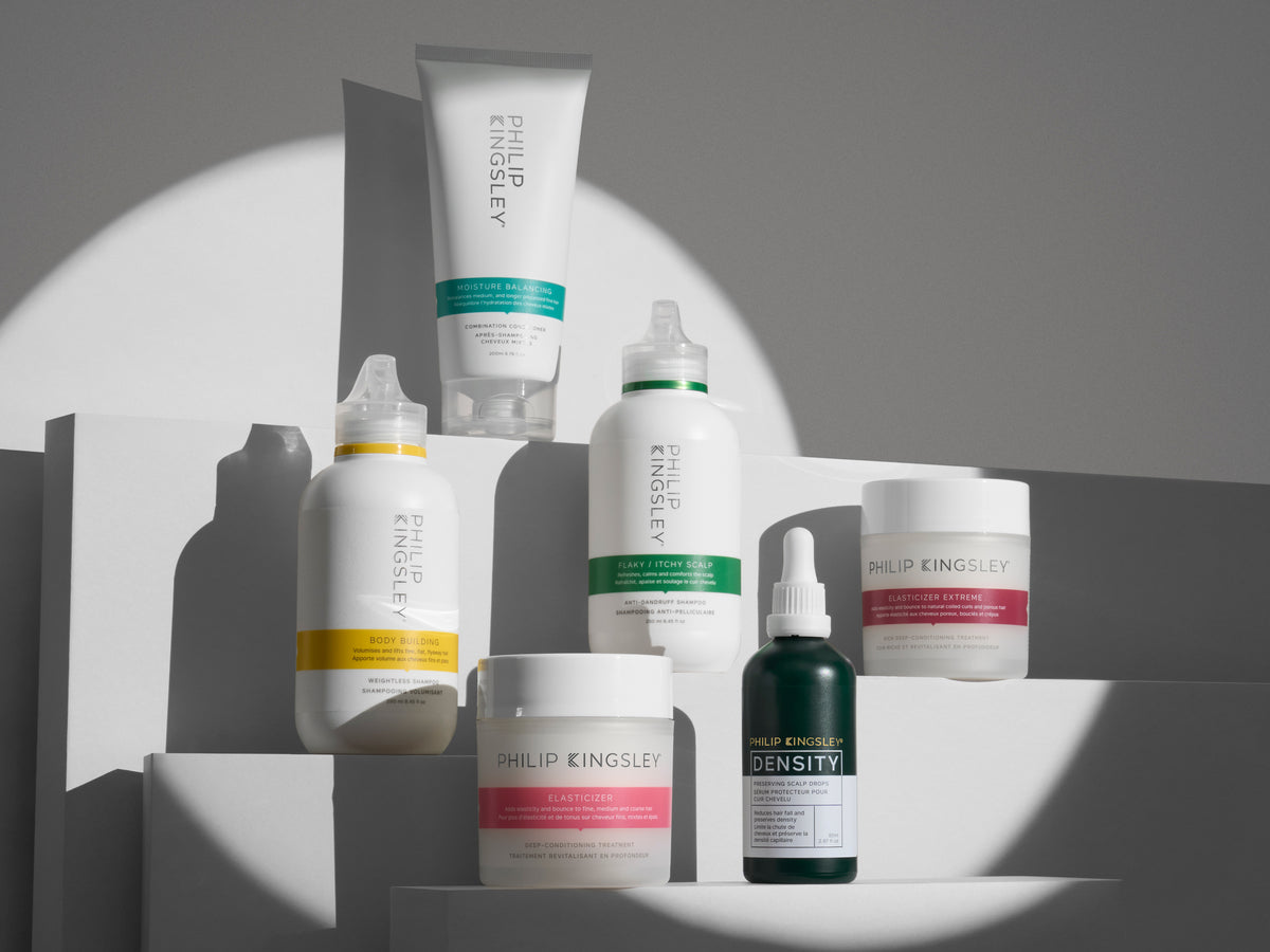Philip Kingsley hair care range in white packaging with coloured bands, including Elasticizer, a scalp toner, masks and Density drops, arranged on white steps in a spotlight