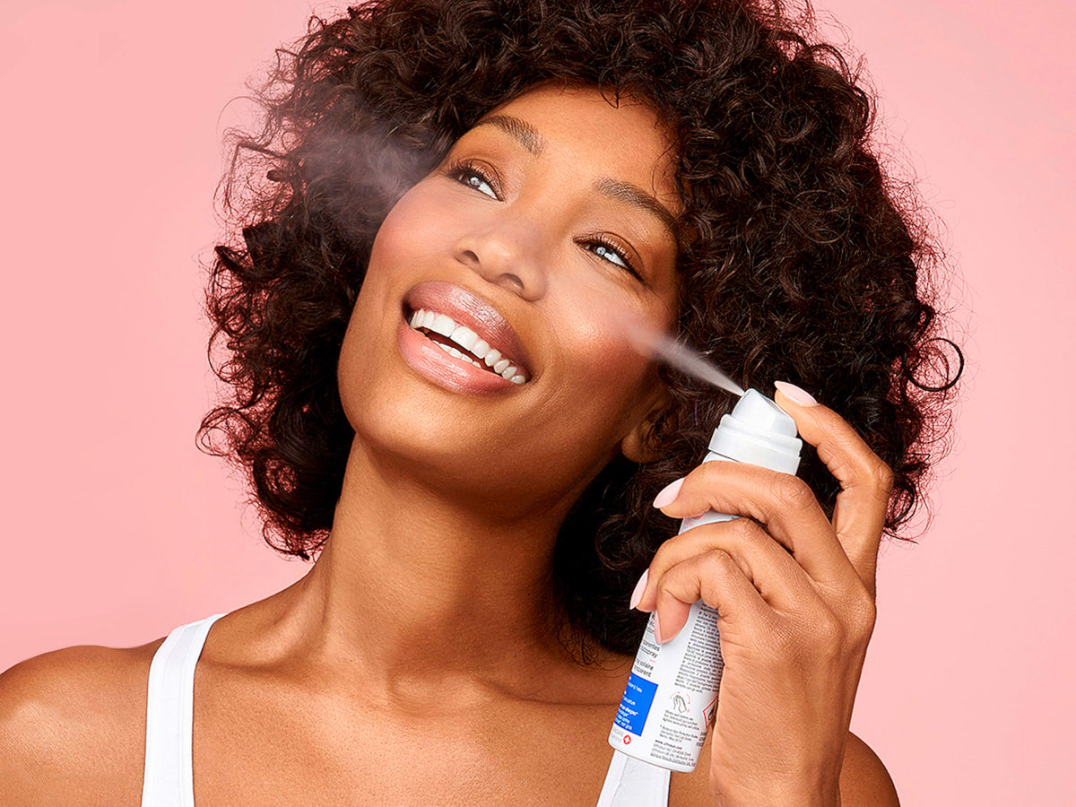 Smiling woman with curly dark hair spraying Ultrasun Face and Scalp UV Protection Mist SPF50 onto her face, with a fine mist visible, against a soft pink background