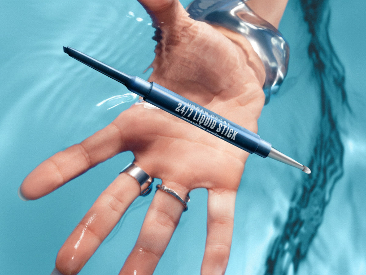 Urban Decay 24/7 Liquid Stick Retractable Eyeliner in a slim blue pen, lying across a hand with silver rings and a silver cuff, beneath rippling turquoise water